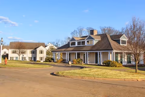 Exterior view of a senior living facility with two buildings surrounded by grass and trees under a clear blue sky. The building on the right has a large covered porch with white columns and dormer windows on the roof. The building on the left is a two-story structure with multiple windows.
