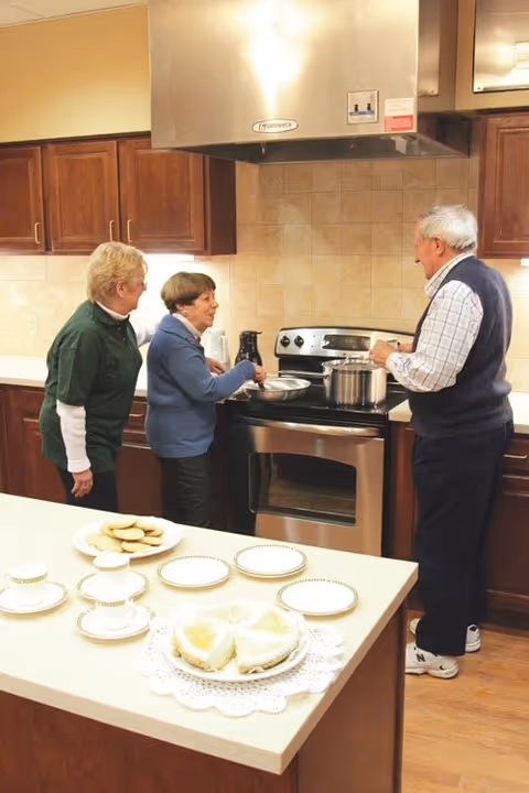 Three older adults standing around a kitchen stove preparing food while plates and desserts sit on the island countertop.