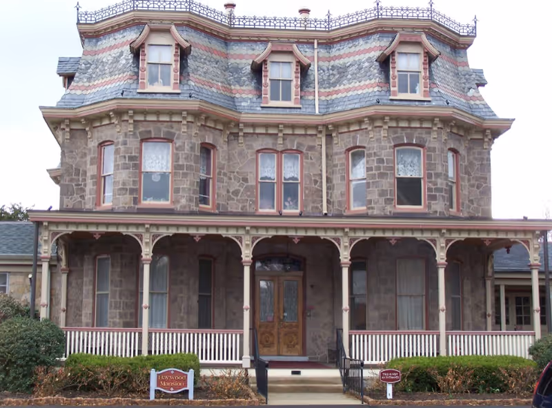 Front exterior view of a large, historic stone mansion with a covered porch, ornate wooden double doors, and multiple windows with lace curtains. The roof has decorative shingles and ironwork along the top edge. There are bushes and a small sign in front of the building.