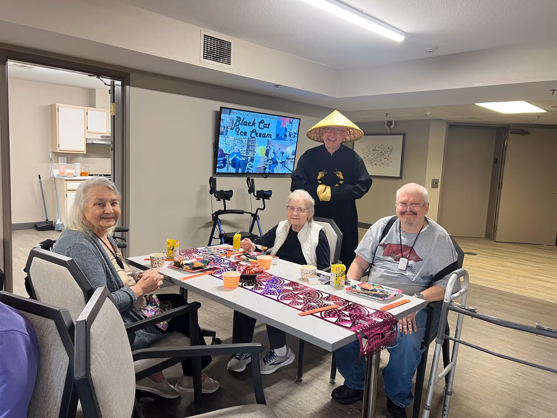 Three elderly people sitting around a table with food and drinks in a common area of a senior living facility. A person dressed in a traditional Asian outfit and hat stands behind them smiling. A television screen on the wall displays the text 'Black Cat Ice Cream'. The room has light-colored walls and flooring, with an open doorway leading to a kitchen area.