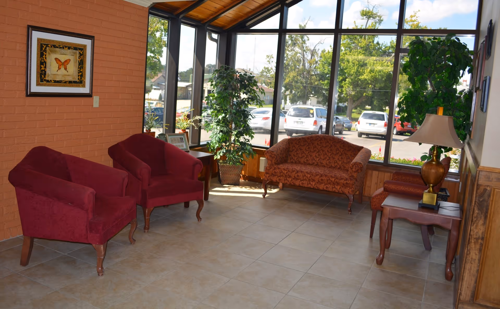 A bright sitting area with large floor-to-ceiling windows showing a parking lot outside. The room has two red armchairs, a patterned red loveseat, a wooden side table with a lamp, a potted plant, and a framed butterfly picture on a brick wall.