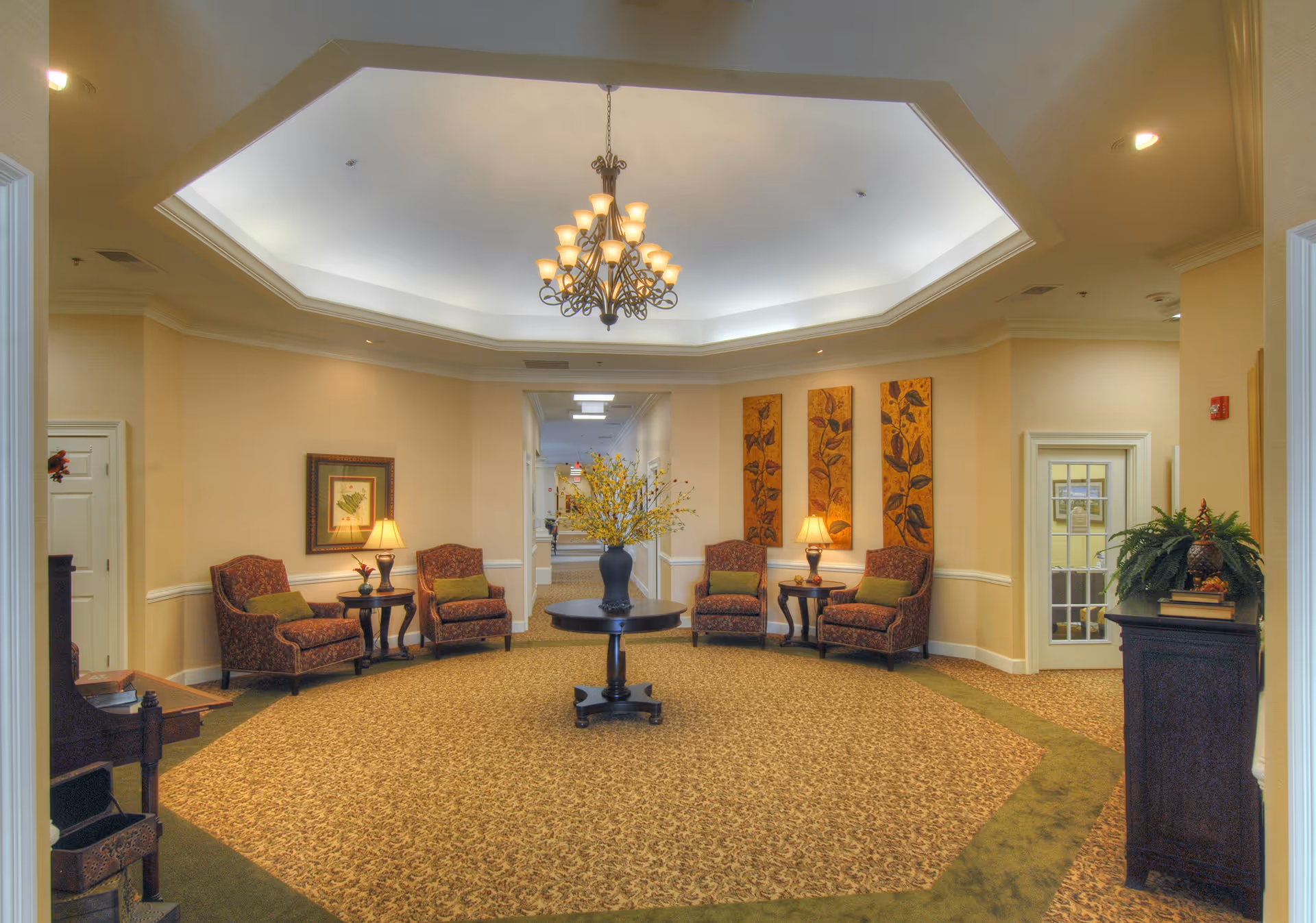 A spacious and elegantly decorated common area with a large octagonal recessed ceiling featuring a chandelier. The room has patterned carpet with a green border, four upholstered armchairs with green cushions arranged around two small side tables, and a round table in the center with a vase of yellow flowers. The walls are adorned with framed artwork and three vertical floral panels. There is a hallway visible in the background and a door with glass panes on the right side.