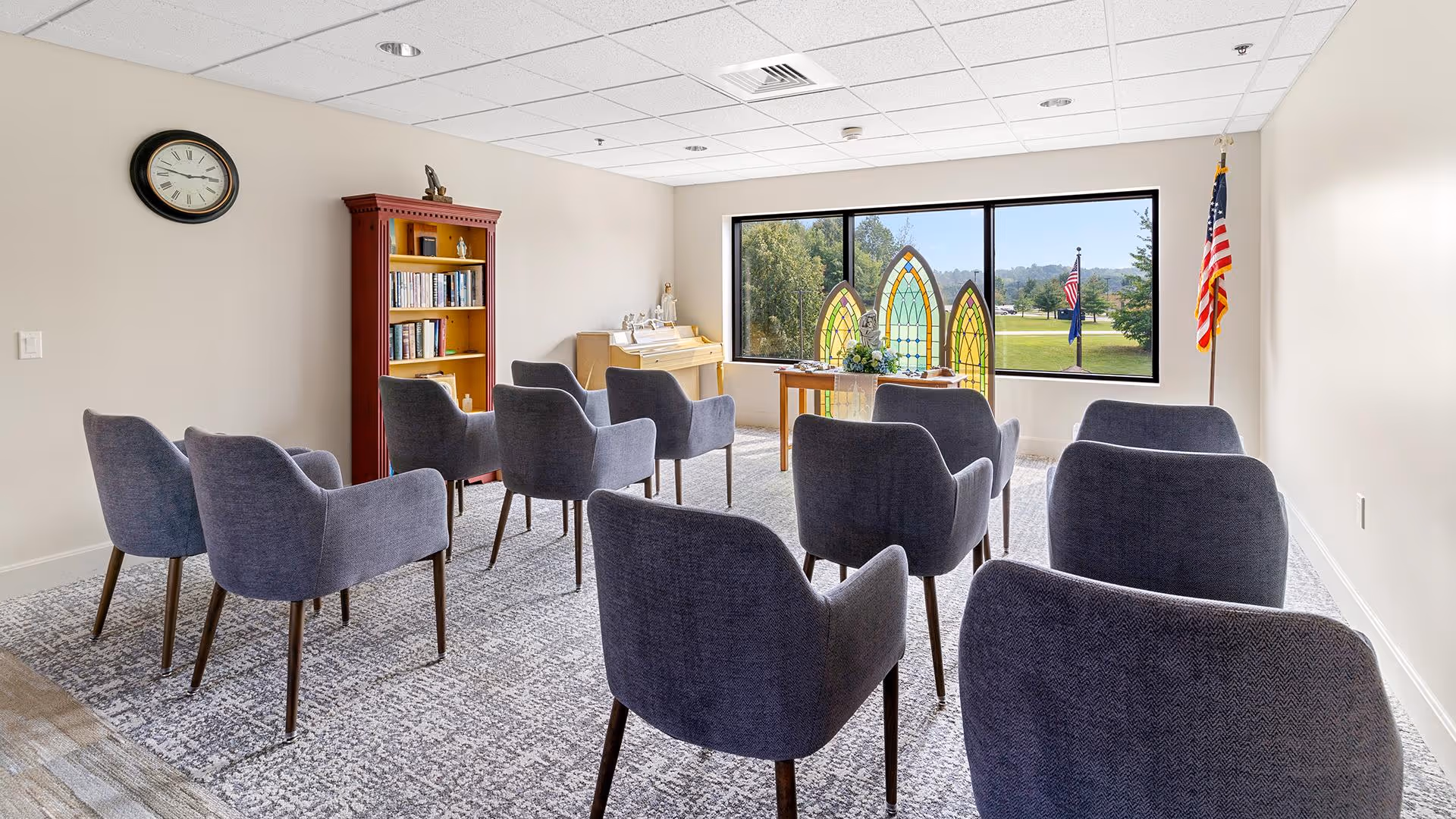 A small meeting or chapel room with two rows of gray upholstered chairs facing a table with decorative stained glass panels in front of a large window. There is a wooden bookshelf with books and a clock on the wall. An American flag stands in the corner near the window.