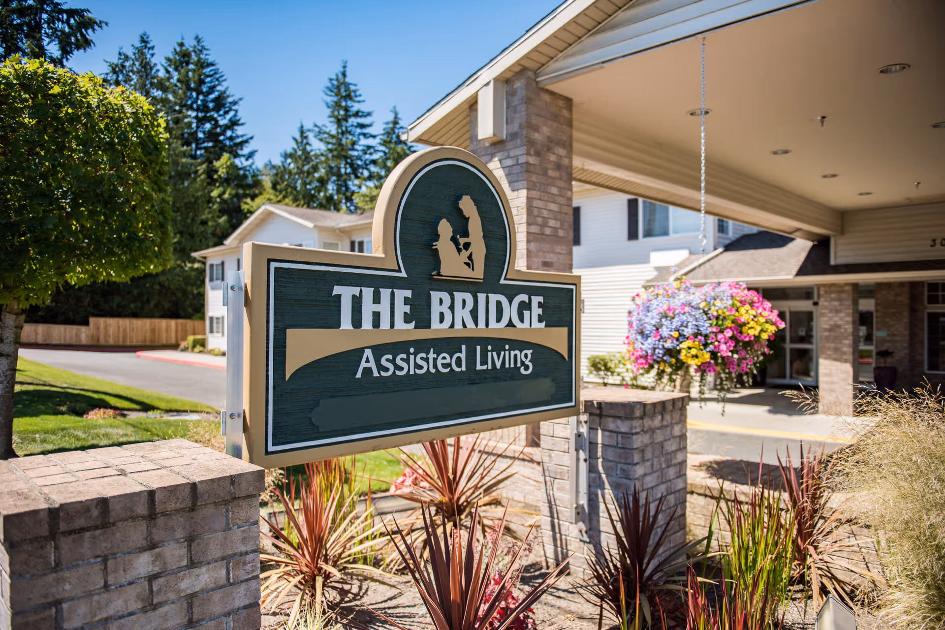 Entrance sign reading "The Bridge Assisted Living" in front of the facility with hanging flowers and a covered driveway.