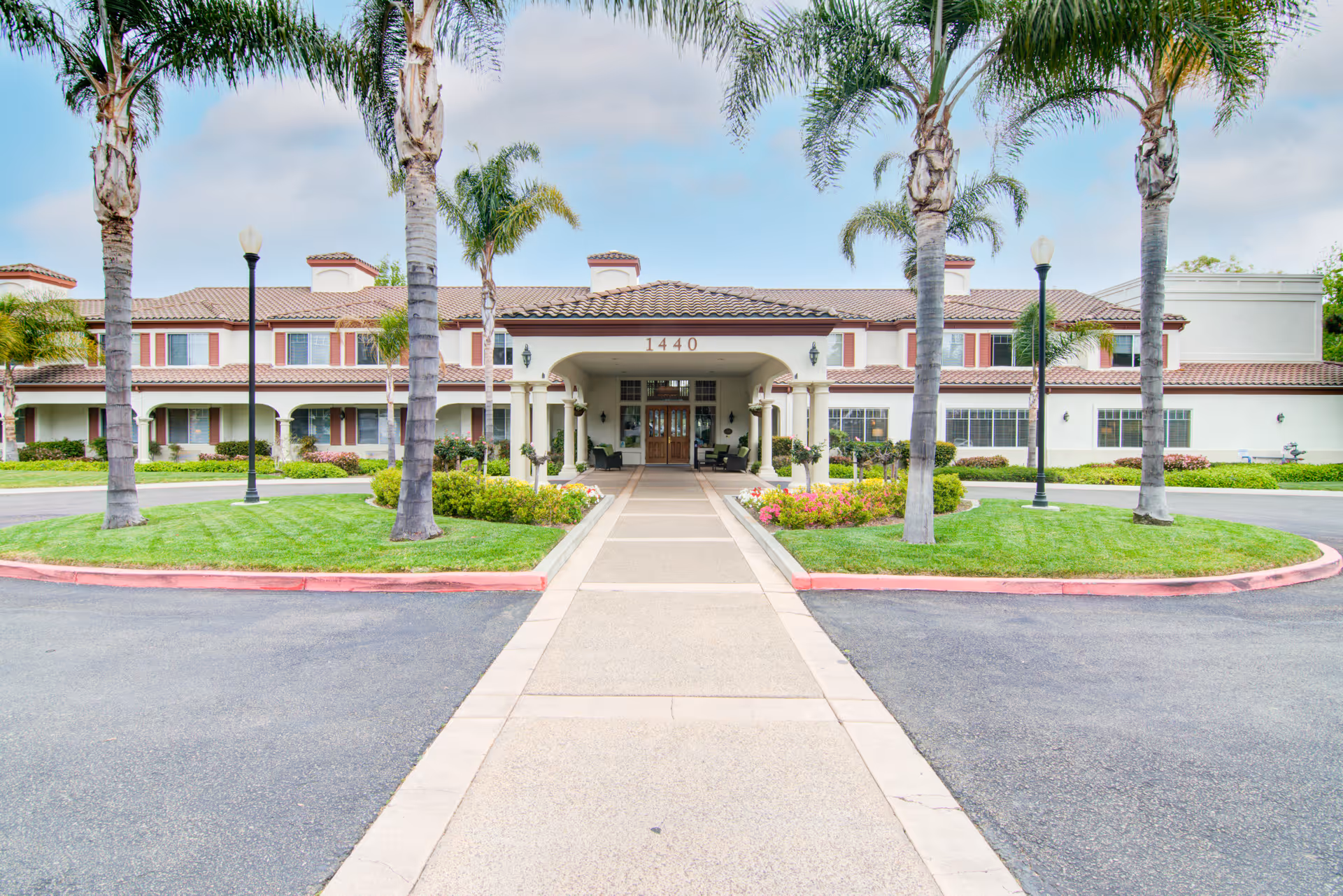Front exterior view of a two-story senior living facility with a tiled roof, palm trees, green lawns, and a central walkway leading to the main entrance under a covered porch. The building number 1440 is displayed above the entrance.