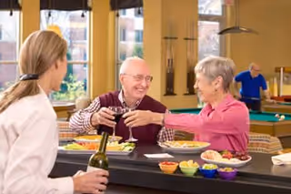 Two elderly residents clinking wine glasses at a dining counter while a staff member pours wine and another person plays pool in the background.