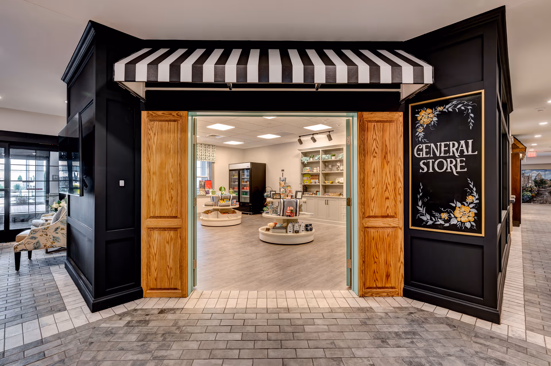 Entrance to a general store inside a senior living facility, featuring a black and white striped awning, wooden double doors, and a black wall with a floral sign that reads 'GENERAL STORE'. Inside, shelves and display tables hold various items for sale.
