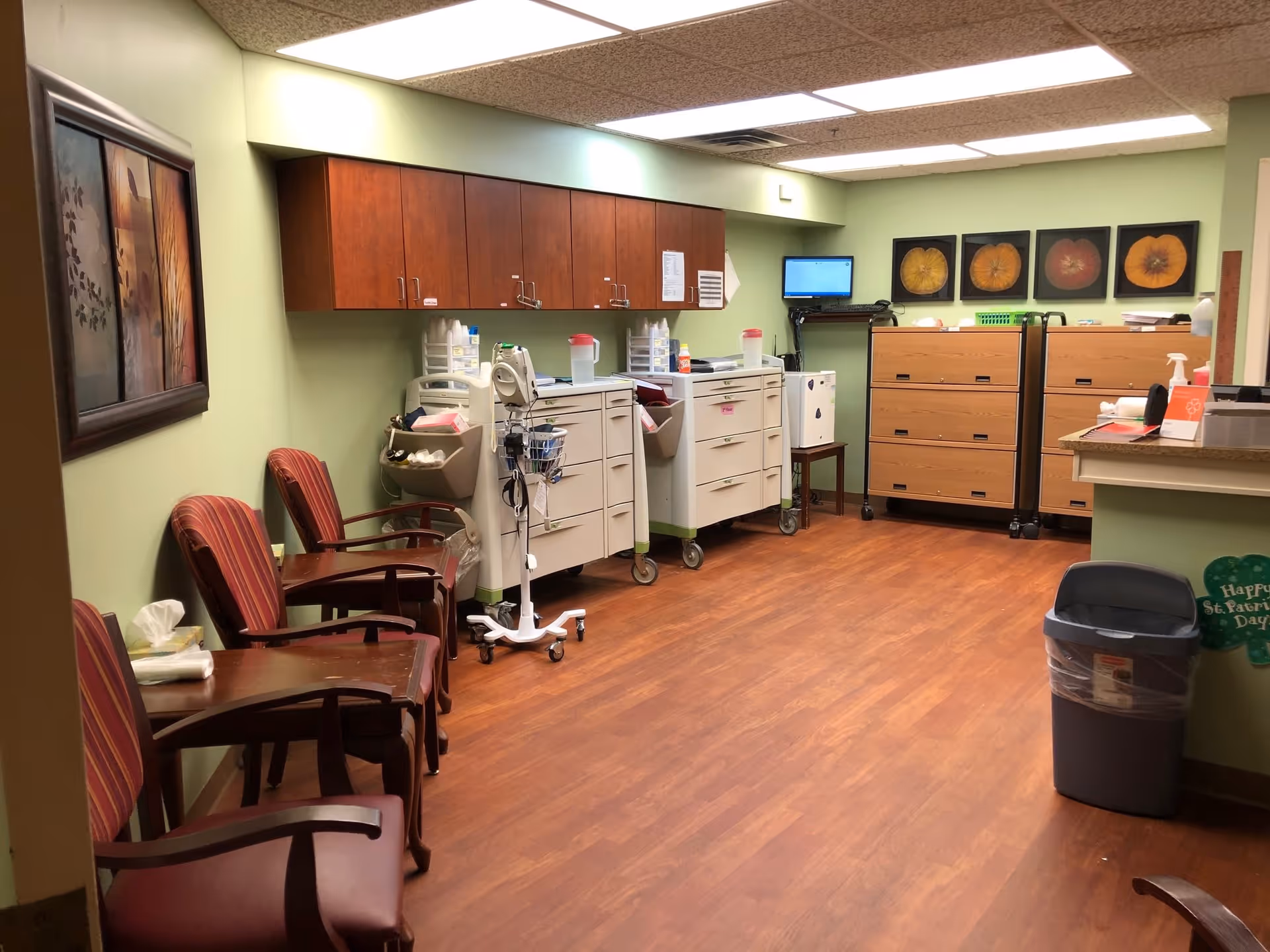 Interior room with wooden floor and light green walls, featuring several red cushioned chairs with wooden armrests lined up against the left wall. On the right side, there are medical carts with supplies, a small monitor mounted on the wall, and wooden filing cabinets. The room is well-lit with ceiling lights and decorated with framed pictures on the walls.
