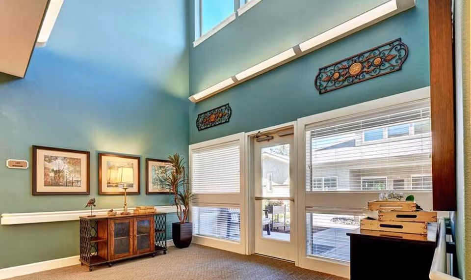 A bright interior room with teal walls and large windows letting in natural light. The room features a wooden cabinet with a lamp and decorative items, three framed pictures on the wall, a tall potted plant, and decorative wall hangings above the windows and door. The door leads outside to a patio area.