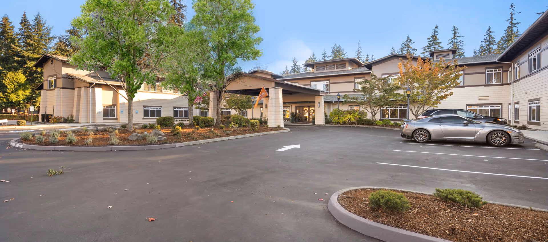 Front entrance and driveway of a two-story senior living facility with landscaping, trees, and a parked car.