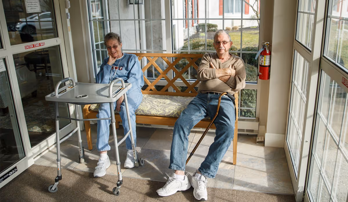 Two elderly residents sit on a wooden bench in a sunlit lobby by large windows, with a walker and a cane nearby.