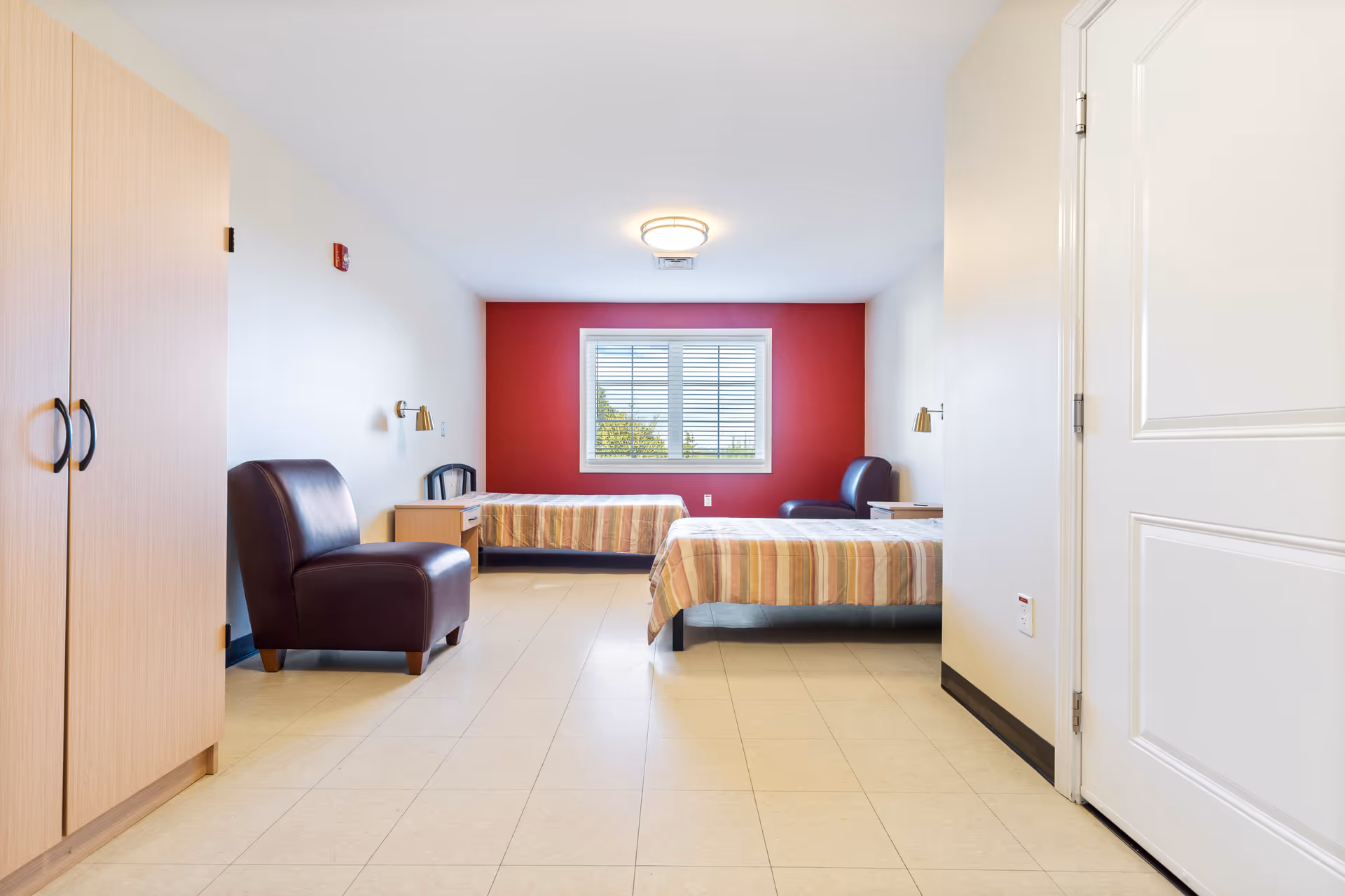 A simple, clean bedroom with two single beds covered in striped bedspreads, two dark brown leather chairs, a wooden wardrobe, two small wooden nightstands, and a window with white blinds on a red accent wall.