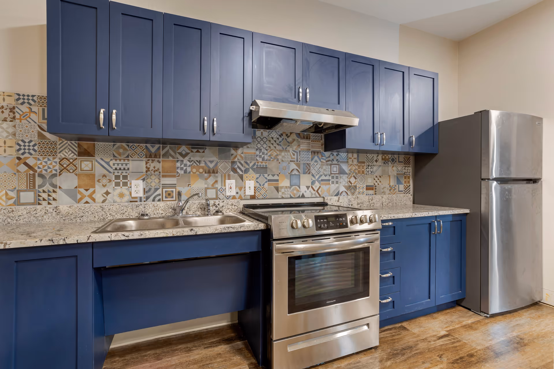 Modern kitchen with blue cabinets, a stainless steel refrigerator, stove, and range hood. The backsplash features a colorful patterned tile design, and the countertop is a light granite. The floor is wood-style.