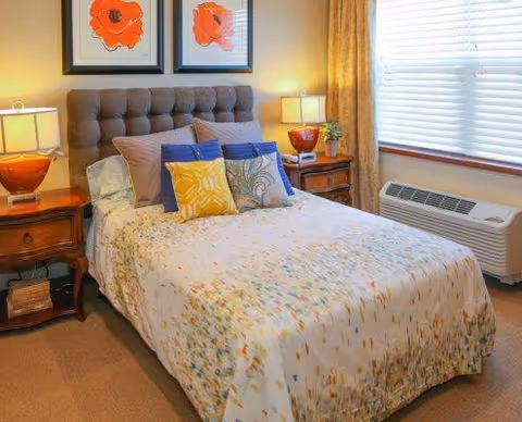 A cozy bedroom with a neatly made bed featuring a floral-patterned bedspread and multiple decorative pillows in blue, yellow, and beige. The bed has a tufted headboard and is flanked by two wooden nightstands, each with a lamp. Above the bed, there are two framed floral artworks. A window with closed blinds and beige curtains is on the right side, and below it is a wall-mounted air conditioning unit.
