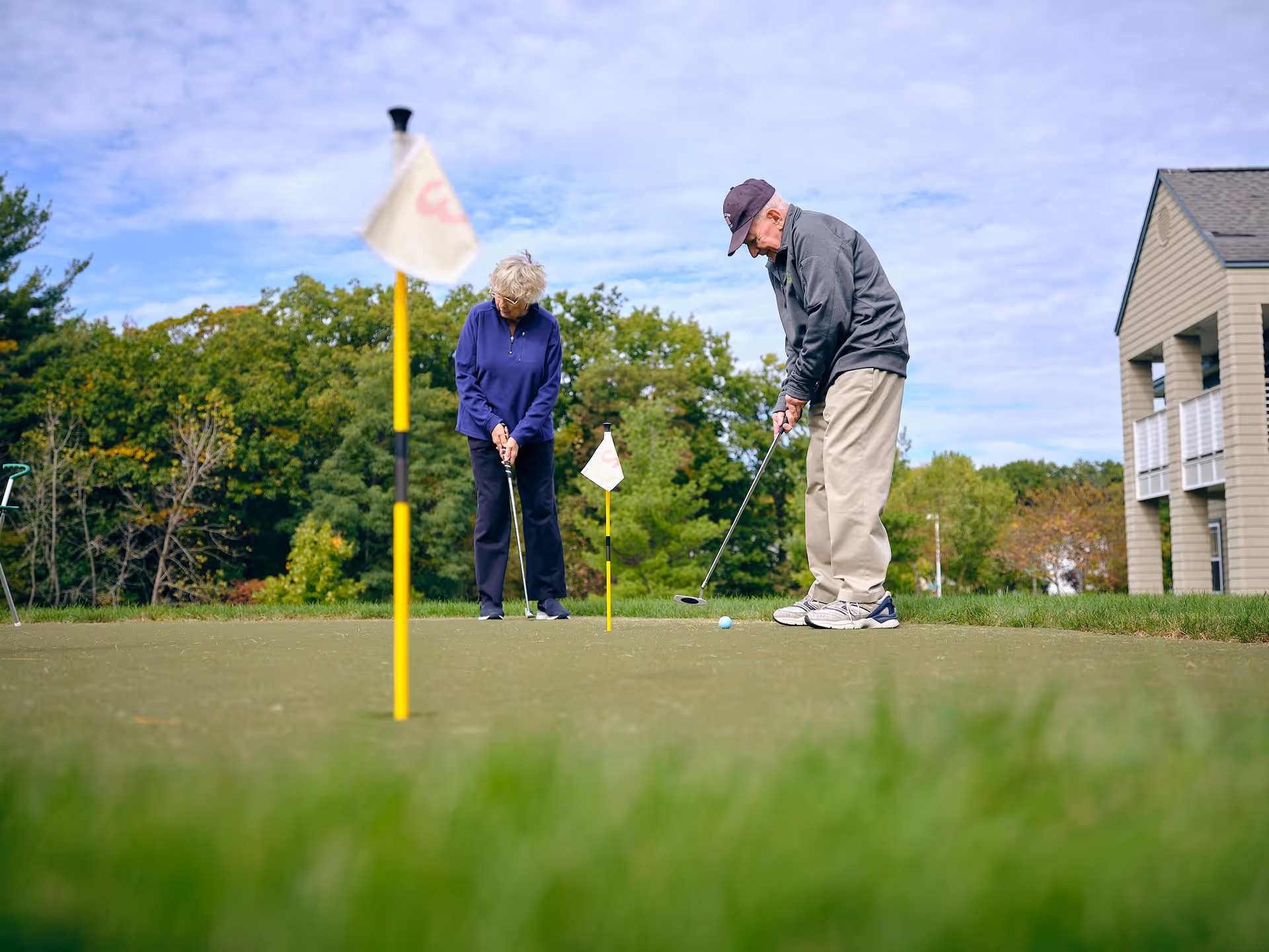 Two elderly people playing golf on a putting green outdoors near a building, with trees and a partly cloudy sky in the background.