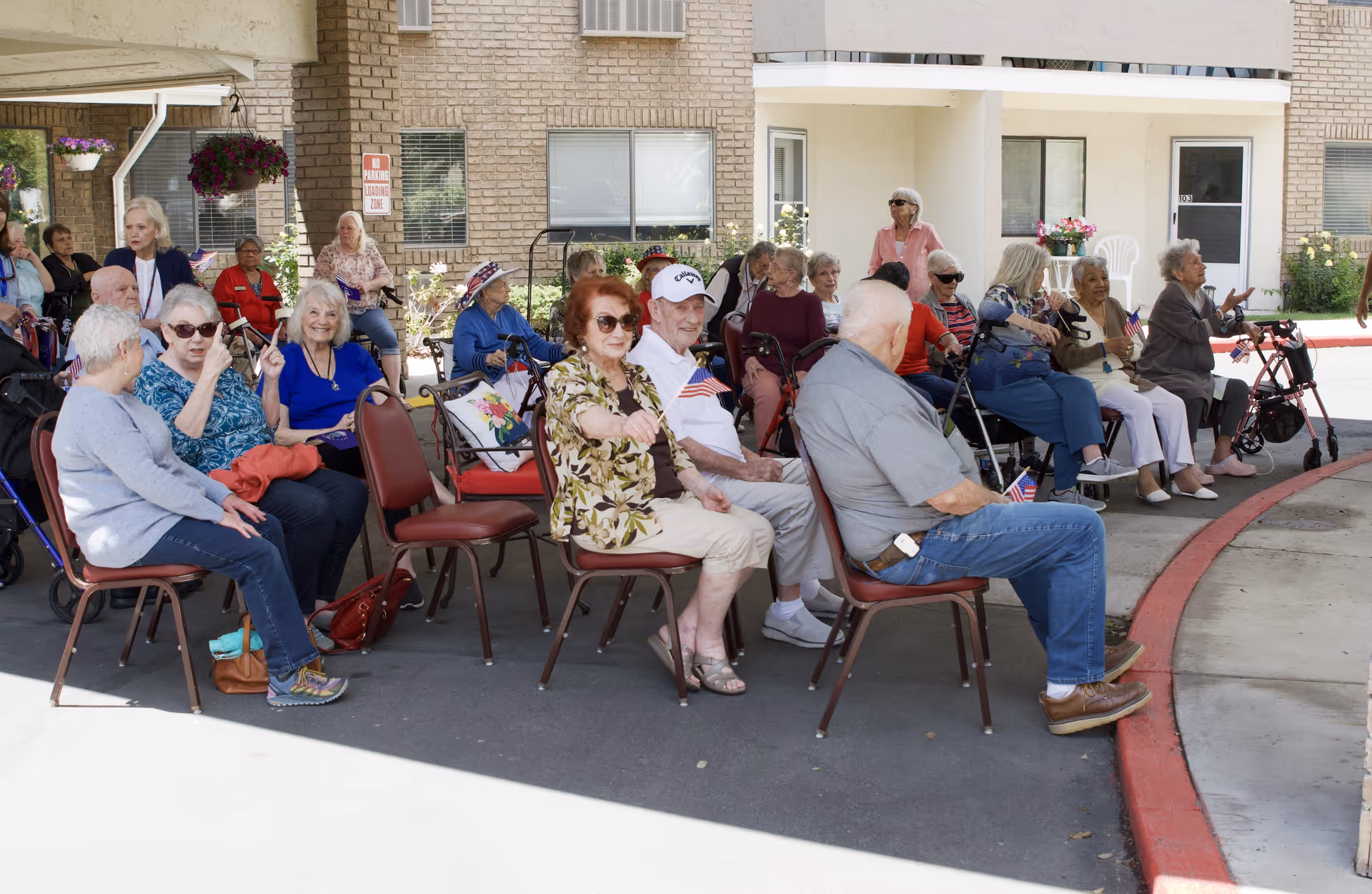 A group of elderly people sitting outdoors on chairs and some in wheelchairs, many holding small American flags, in front of a brick building with windows and doors. Some are engaged in conversation, and the setting appears to be a social or community event at a senior living facility.