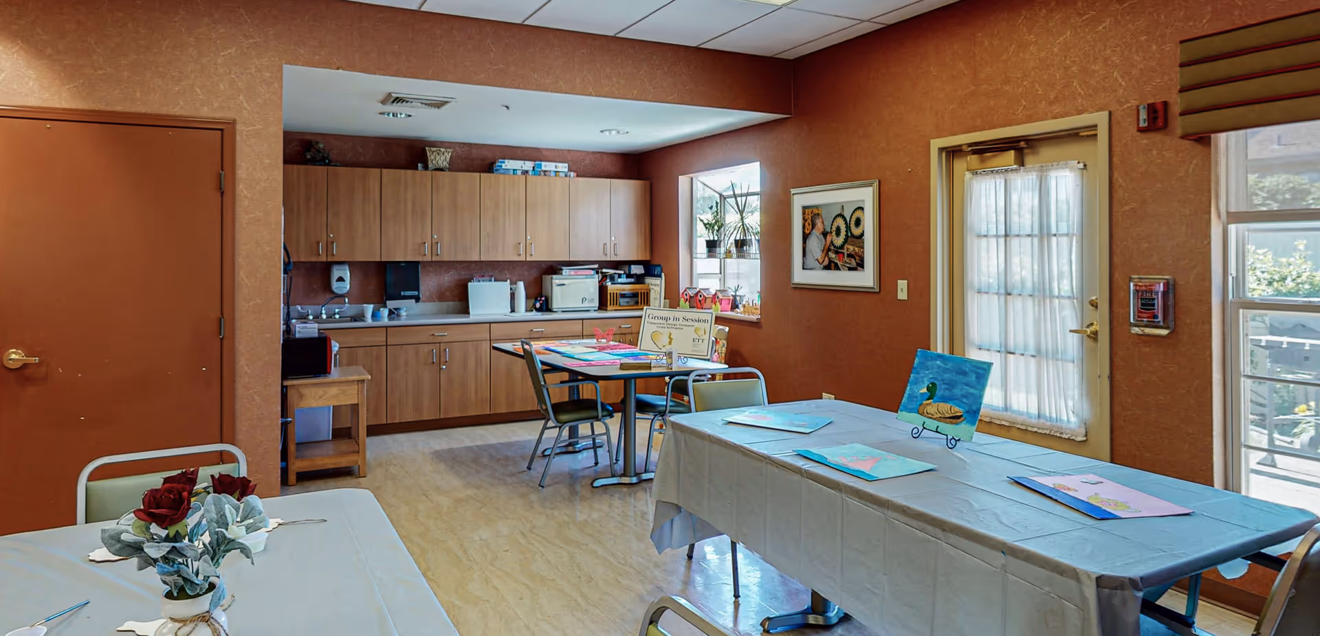A well-lit room with tables covered in white tablecloths, some with art supplies and paintings on them. The room has wooden cabinets along one wall with a sink and various items on the countertop. There are chairs around the tables, a door with a window covered by a curtain, and a window letting in natural light. The walls are painted a warm brown color, and there is a framed picture hanging on one wall.