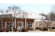 Exterior view of a single-story brick building with white trim and a covered entrance supported by columns. There are leafless trees and shrubs in front of the building under a clear sky.