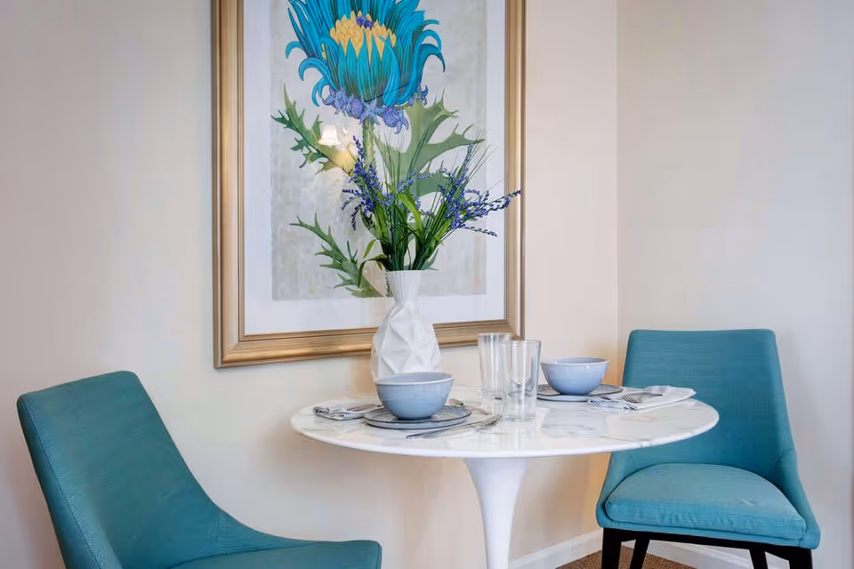 Small dining nook with a round white table set for two, teal chairs, a vase of flowers, and framed floral artwork on the wall.