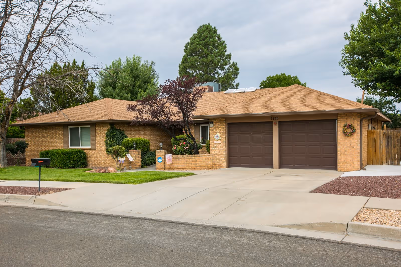 Single-story brick building with a brown shingled roof, two closed brown garage doors, a small front yard with green grass, bushes, and trees, and a concrete driveway under a cloudy sky.