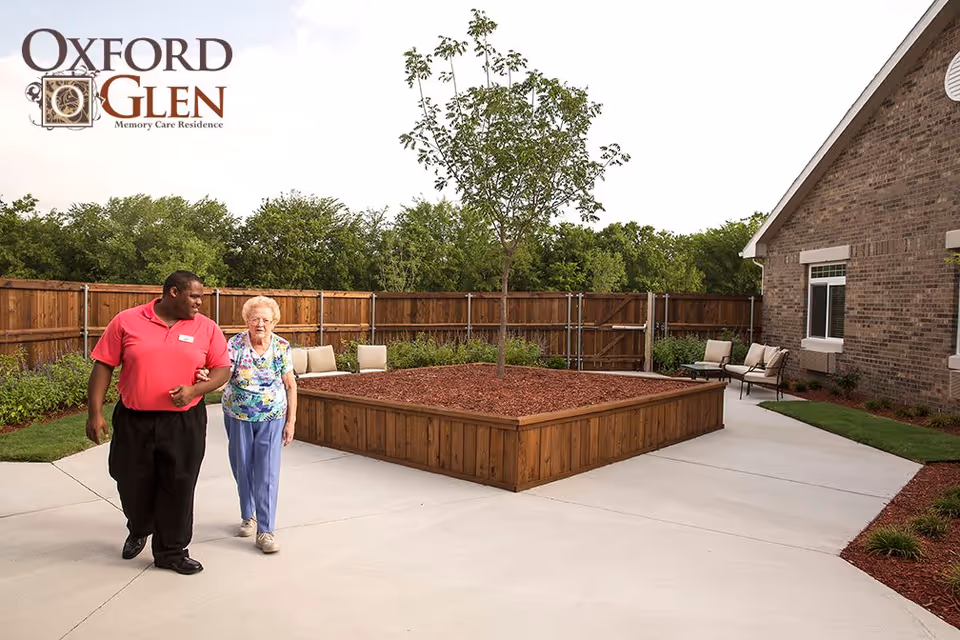 An outdoor courtyard area at Oxford Glen Memory Care Residence with a raised wooden planter containing a small tree in the center. A man in a red shirt is walking arm-in-arm with an elderly woman wearing a floral blouse and blue pants. There are chairs along the wooden fence and a brick building on the right side.