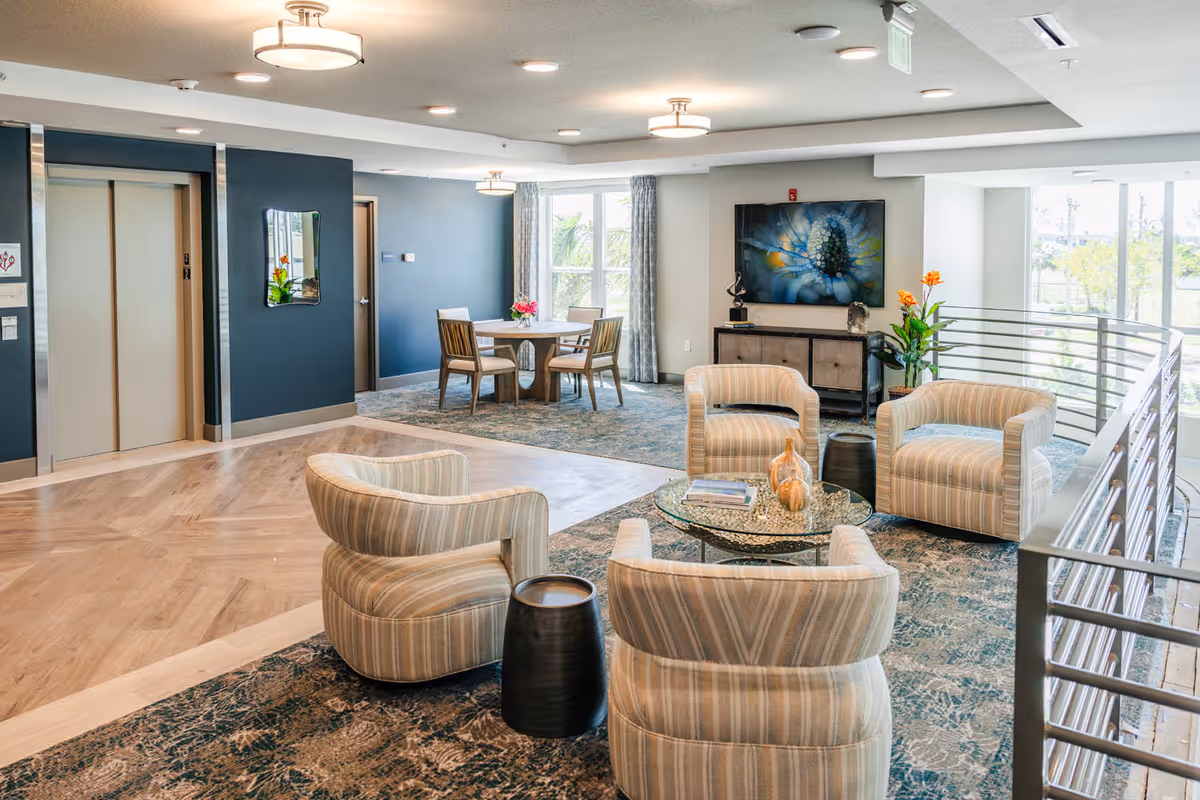 A bright and modern lounge area in a senior living facility featuring four striped armchairs arranged around a glass coffee table with decorative items. In the background, there is a round wooden table with four chairs near large windows with curtains. The walls are painted in blue and white tones, and there are two elevators on the left side. The space is well-lit with ceiling lights and natural light from the windows.