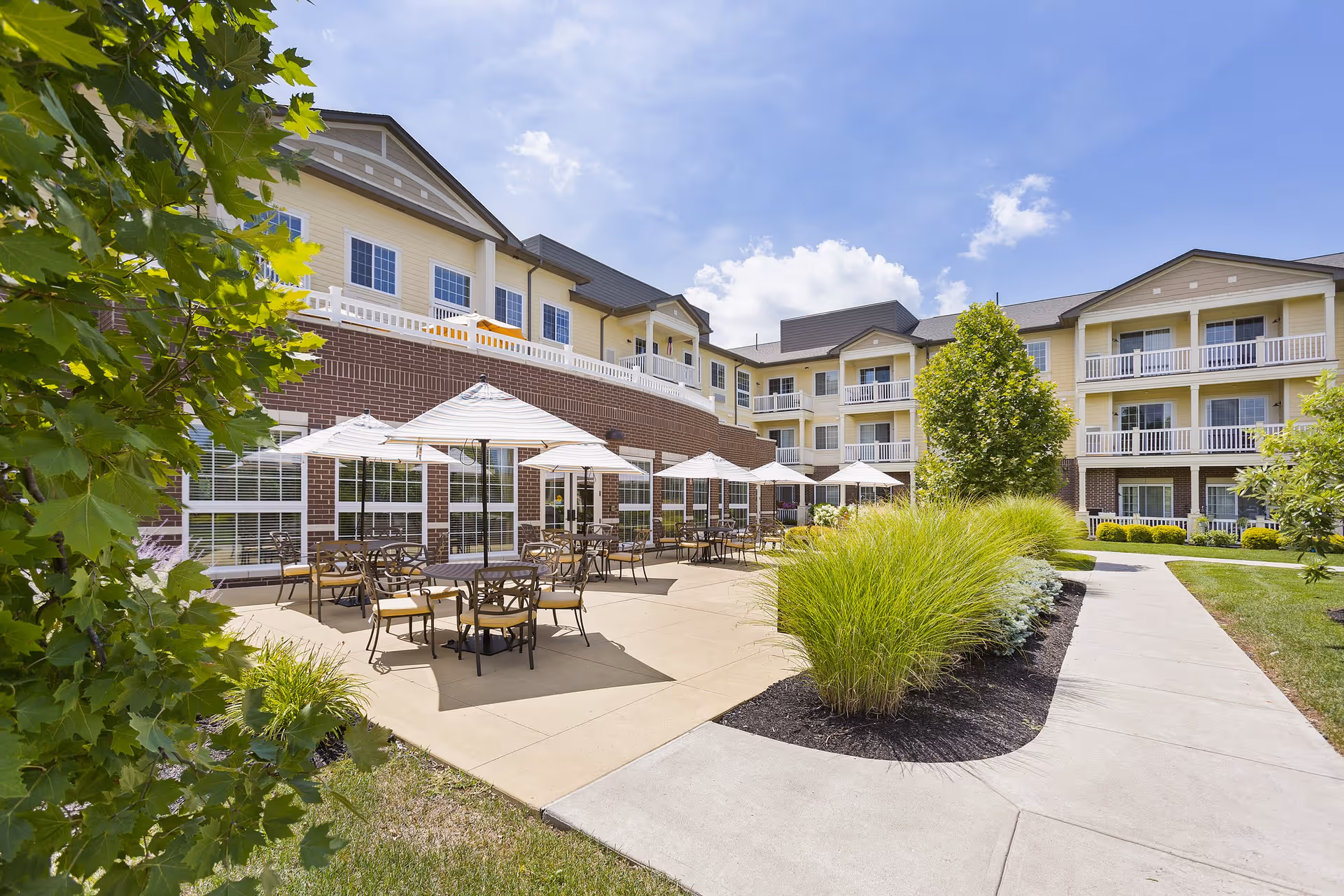 Outdoor patio area of a senior living facility with multiple tables and chairs under white umbrellas, surrounded by greenery and a three-story building with balconies in the background under a blue sky with some clouds.