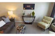 Small living room with a gray sofa, patterned armchair, wall-mounted TV, glass-top coffee table, and a console table with decorative items.