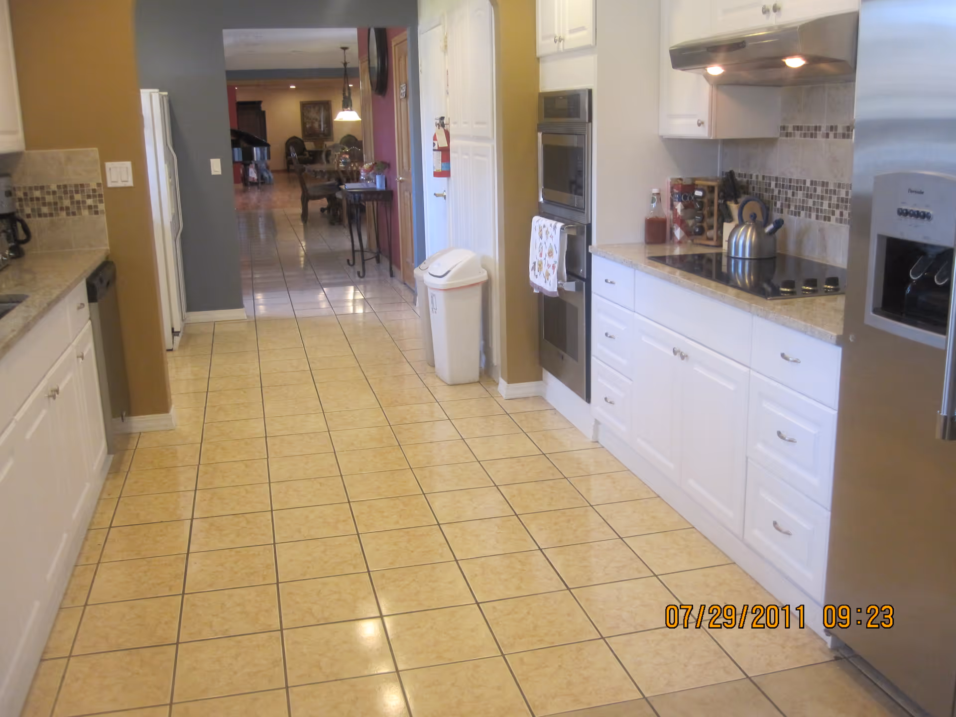A clean, modern kitchen with white cabinets, beige tiled floor, and stainless steel appliances including a refrigerator and built-in oven. The kitchen has a tiled backsplash with mosaic accents and a kettle on the stovetop. The view extends into a dining area with a table and chairs in the background.