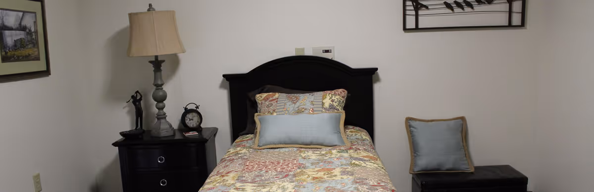 A small bedroom with a single bed featuring a colorful patchwork quilt and two pillows. To the left of the bed is a dark wooden nightstand with a lamp, a small statue, and an alarm clock. On the right side, there is a cushioned bench with a blue pillow. The walls are plain white with a framed picture on the left and a decorative metal wall art above the bed.