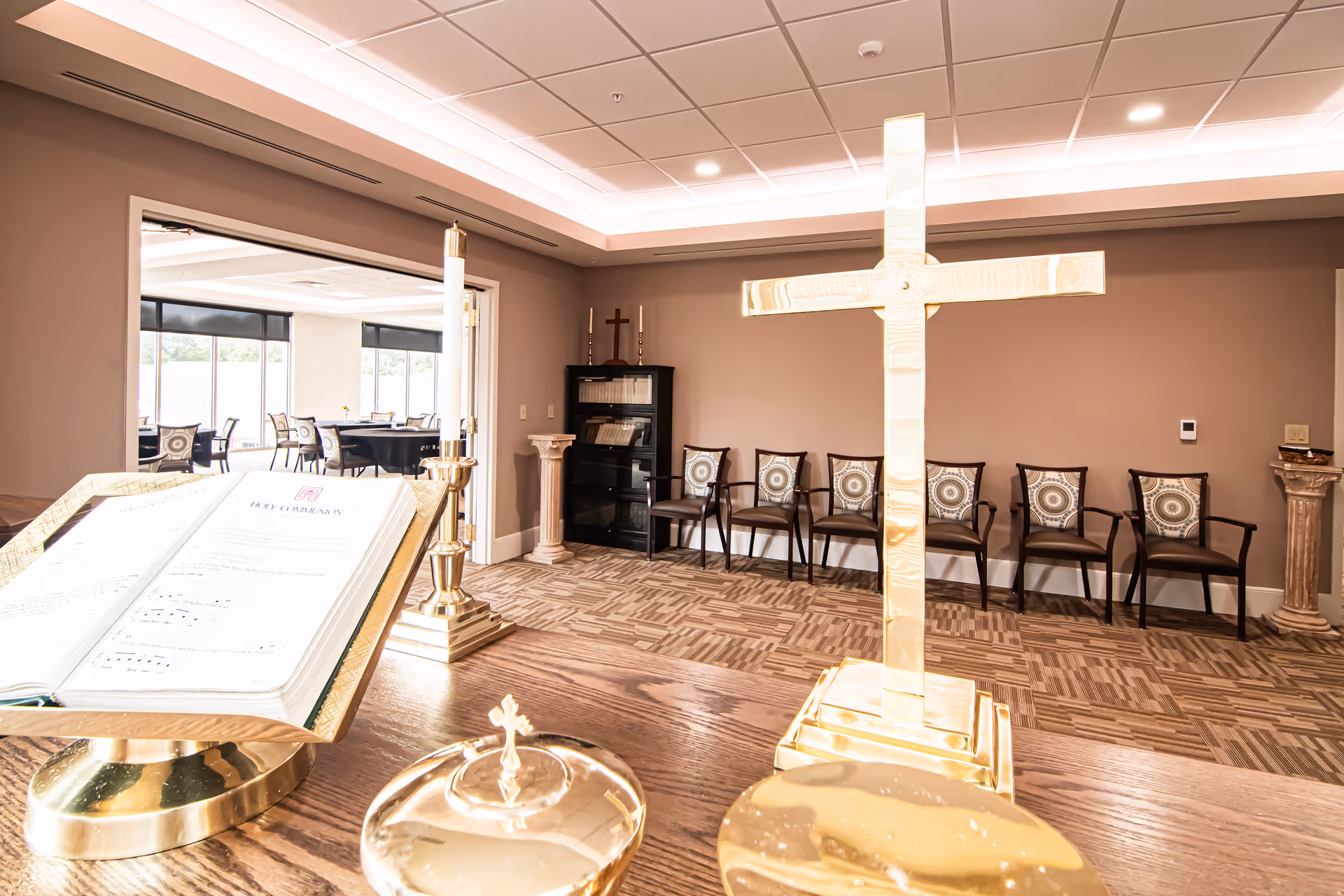 Small interior chapel with a wooden altar featuring a brass cross and hymn book, chairs along the back wall and a dining area visible through an open doorway.