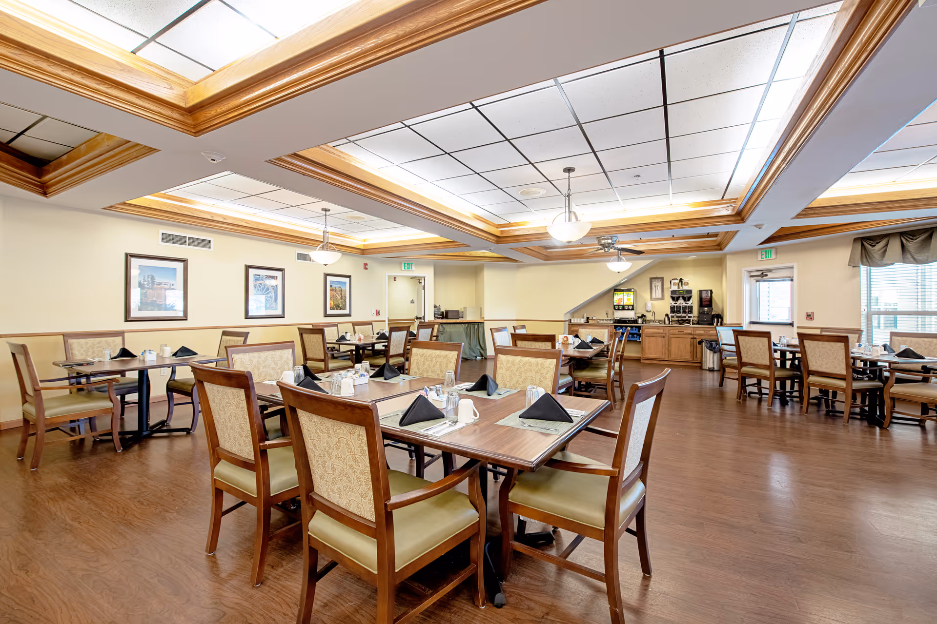 A spacious dining room with multiple wooden tables and chairs arranged neatly. Each table is set with placemats, black folded napkins, cups, and silverware. The room has a wooden floor, cream-colored walls, framed pictures, and a ceiling with recessed lighting and wooden beams. In the background, there is a beverage station with coffee and drink dispensers.