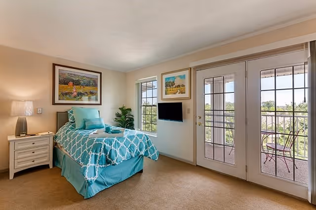 A bright bedroom with a single bed dressed in turquoise and white patterned bedding. There is a nightstand with a lamp beside the bed, two framed paintings on the walls, a small flat-screen TV mounted between two windows, and a glass door leading to a balcony with outdoor seating and a view of greenery.