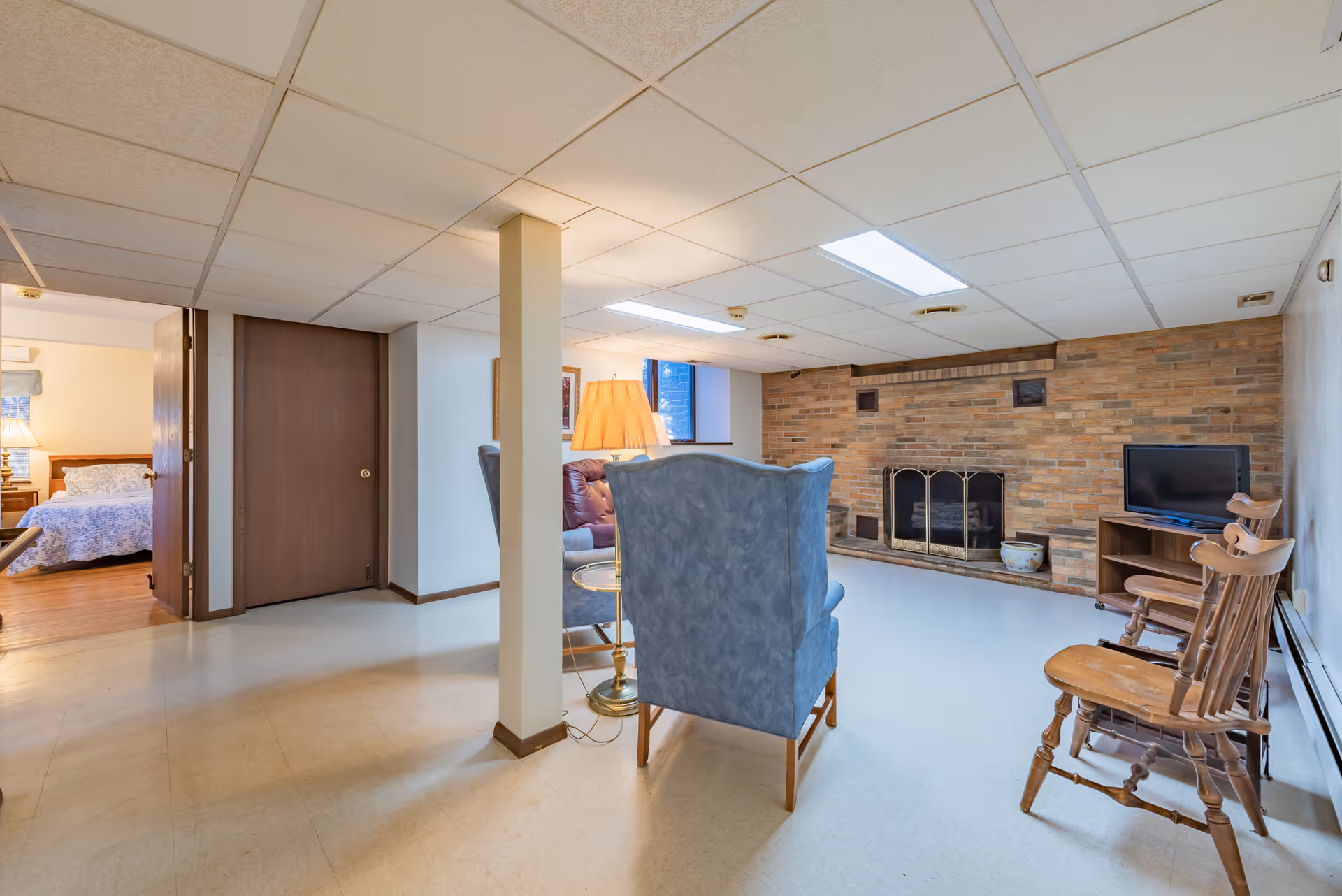 A cozy living room area with a brick fireplace, two blue upholstered armchairs, a small round glass side table with a lamp, and two wooden chairs. A small TV sits on a wooden stand to the right. The room has a drop ceiling with fluorescent lighting and a window letting in natural light. A bedroom with a bed and nightstand is visible through an open door on the left.