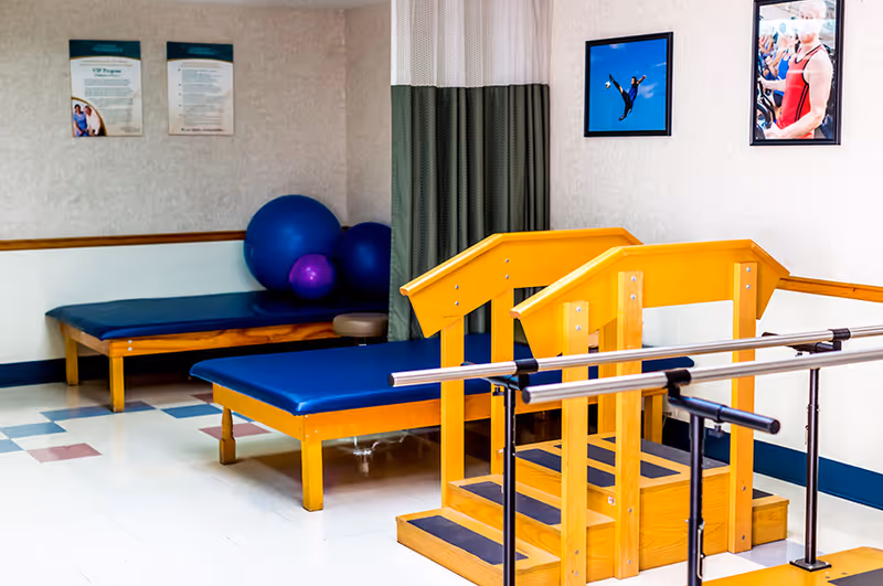 A physical therapy room with two padded treatment tables, several exercise balls, a set of wooden stairs with handrails for rehabilitation, and framed pictures on the walls.
