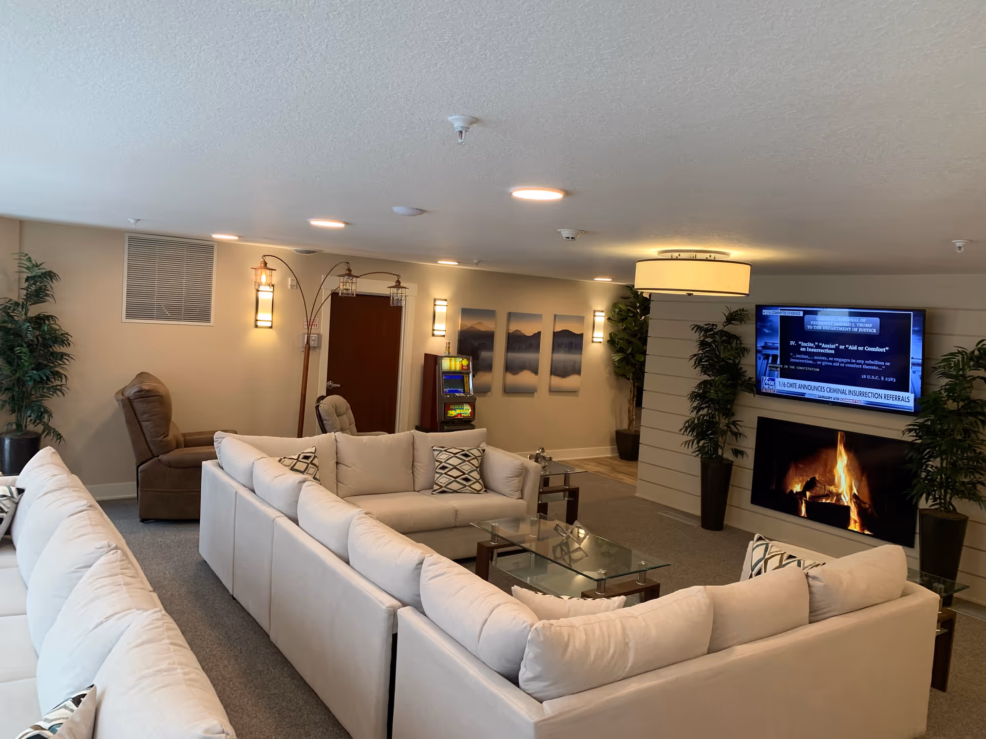 A cozy living room area with beige sectional sofas arranged around a glass coffee table. There is a brown recliner chair in the corner next to a tall floor lamp with three lantern-style lights. On the wall, there are three landscape paintings and a door. A flat-screen TV mounted above a fireplace shows a news broadcast. Two large potted plants flank the fireplace. The room has soft lighting and a neutral color palette.