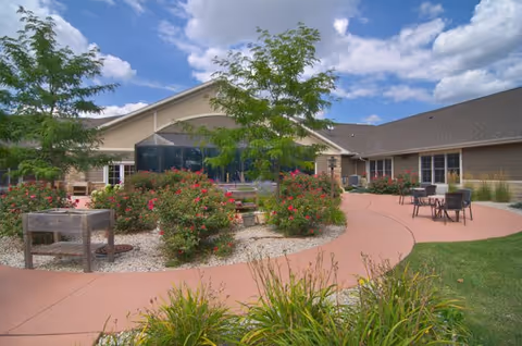 Outdoor patio area at Mission Creek Senior Living featuring a curved concrete walkway surrounded by green grass, flowering bushes, and small trees. There are several tables and chairs for seating, and the building exterior is visible under a partly cloudy blue sky.