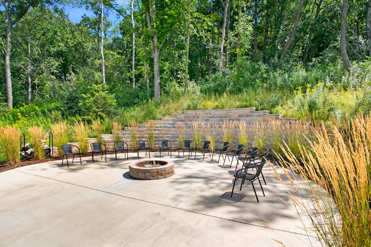 Outdoor seating area with a circular arrangement of black metal chairs around a stone fire pit, surrounded by tall grasses and trees in the background.