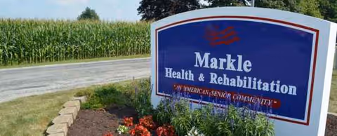 Blue Markle Health and Rehabilitation sign by the roadside with flowers and a cornfield backdrop.