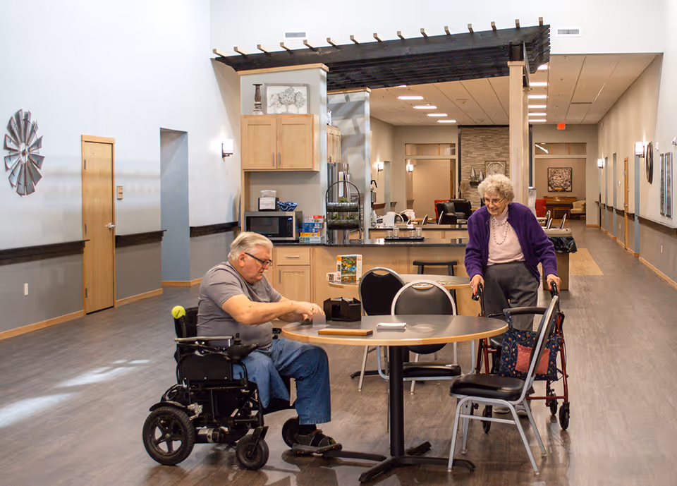 An elderly man in a wheelchair and an elderly woman using a walker are at a round table in a spacious, well-lit common area with wood flooring and light-colored walls. The background shows a kitchen area with cabinets, a microwave, and a countertop, as well as a hallway leading to other rooms.
