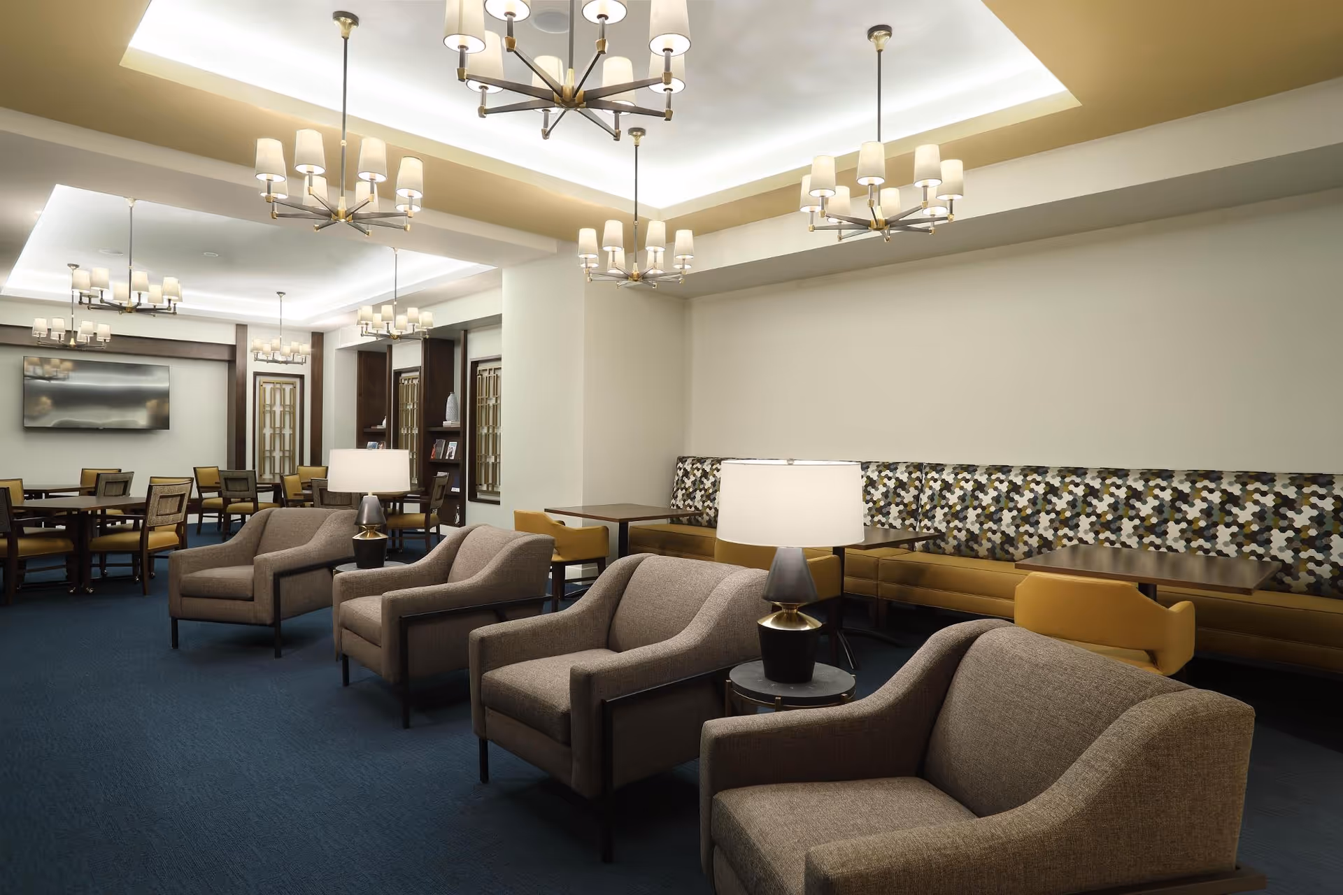 A spacious and well-lit common area with multiple gray armchairs arranged in rows, small round tables with lamps between the chairs, and several dining tables with chairs in the background. The walls are light-colored, and the ceiling features modern chandeliers and recessed lighting. There is a patterned bench along one wall and a large flat-screen TV mounted on the far wall.