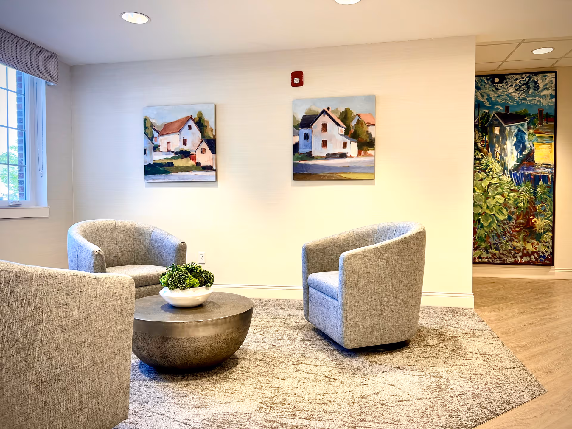 Bright seating area with three gray armchairs around a round coffee table and paintings on the wall.