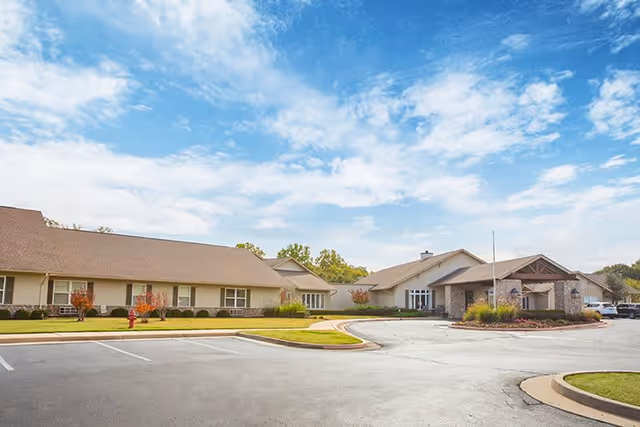 Exterior view of Cedar Ridge Senior Living facility showing a single-story building with a pitched roof, surrounded by a parking lot and landscaped greenery under a partly cloudy blue sky.