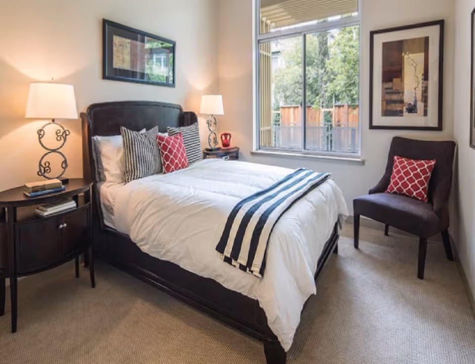A bright bedroom with a neatly made bed, two nightstands with lamps, a window, framed artwork, and an accent chair.