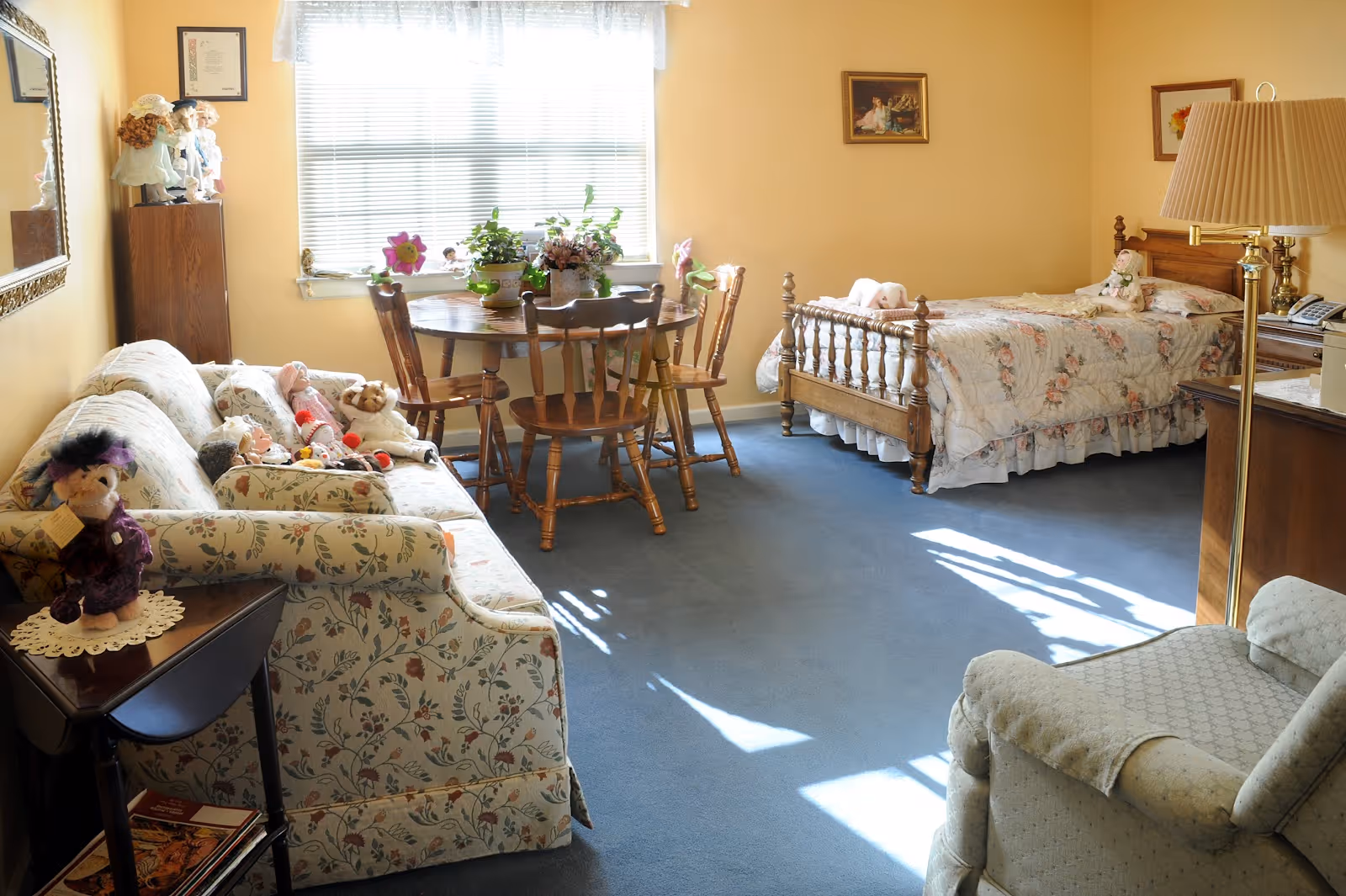 A cozy room with a floral-patterned sofa adorned with stuffed animals, a wooden dining table with four chairs near a window with blinds, a single bed with floral bedding, a wooden nightstand with a lamp, and a comfortable armchair. The room has light yellow walls and blue carpet flooring, decorated with framed pictures and various plants on the table.