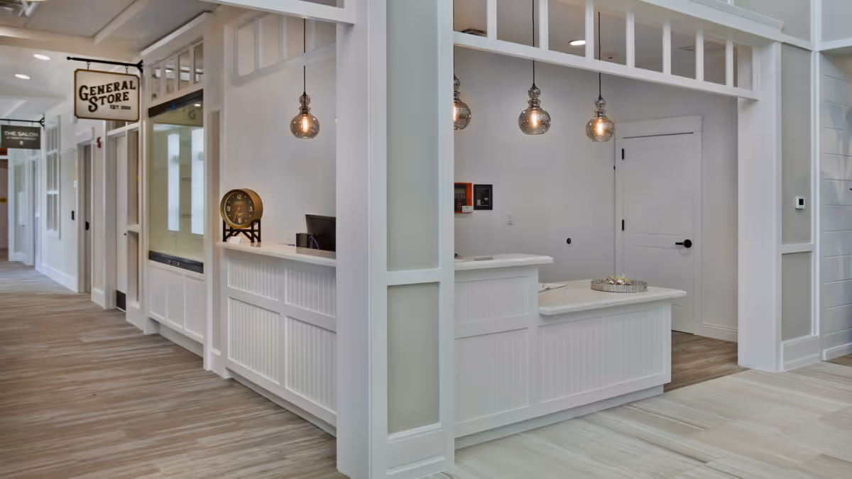 Interior view of a senior living facility hallway with a white reception desk featuring a clock and a computer. Hanging pendant lights illuminate the area. Signs for 'General Store' and 'The Salon' are visible along the corridor.