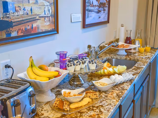 A kitchen countertop with a variety of breakfast items including bananas, yogurt cups, parfaits with blueberries, sliced melon, hard boiled eggs, granola, crackers, and juice pitchers. There is a toaster on the left side and framed artwork on the wall above the counter.