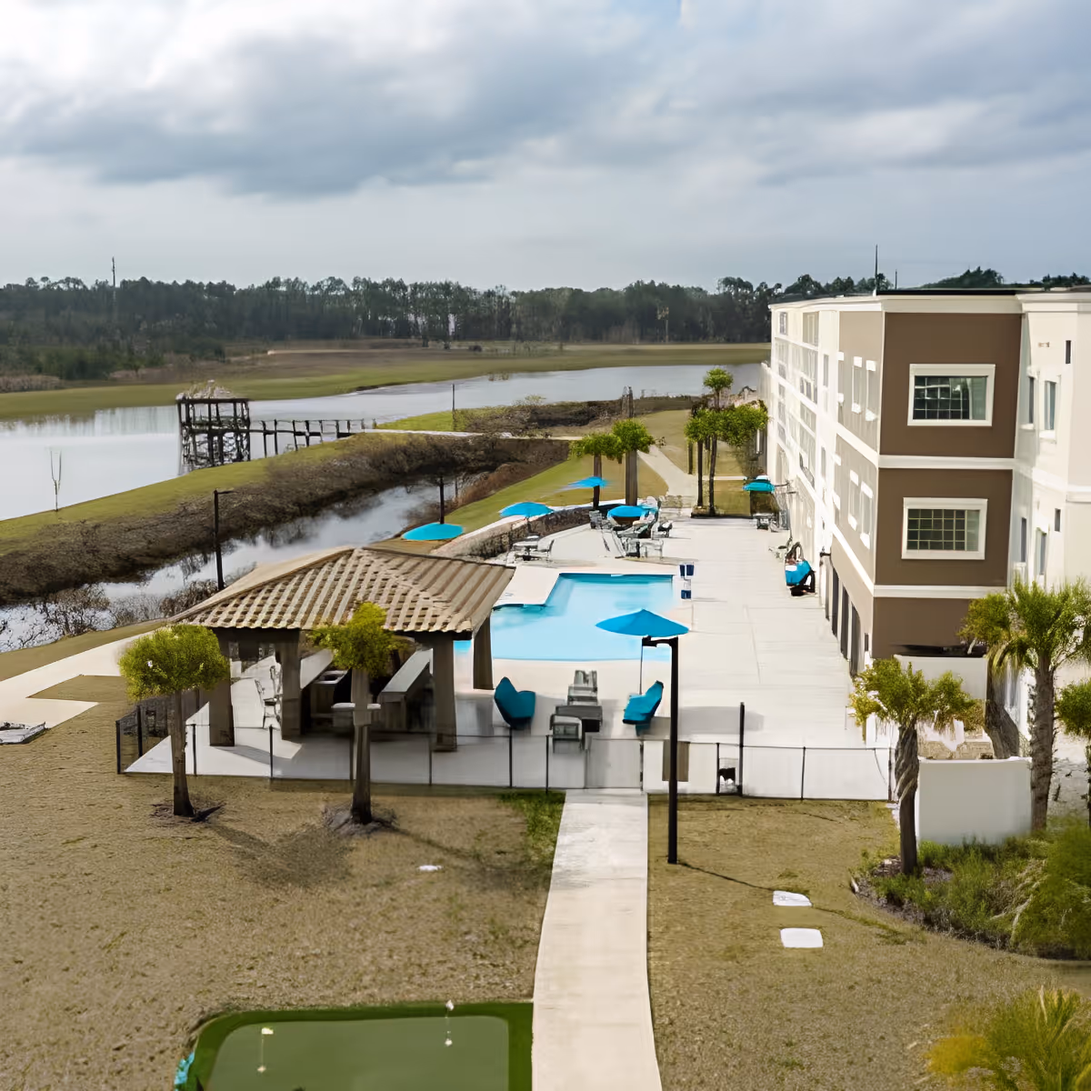 Outdoor view of a senior living facility's pool patio with blue umbrellas and seating next to a multi-story building and a pond in the background.