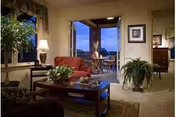 A cozy living room with a red armchair, wooden coffee table with plants and decorative items, a large potted plant on the floor, a lamp on a side table, and a view through an open door leading to an outdoor patio with seating and a scenic evening sky.