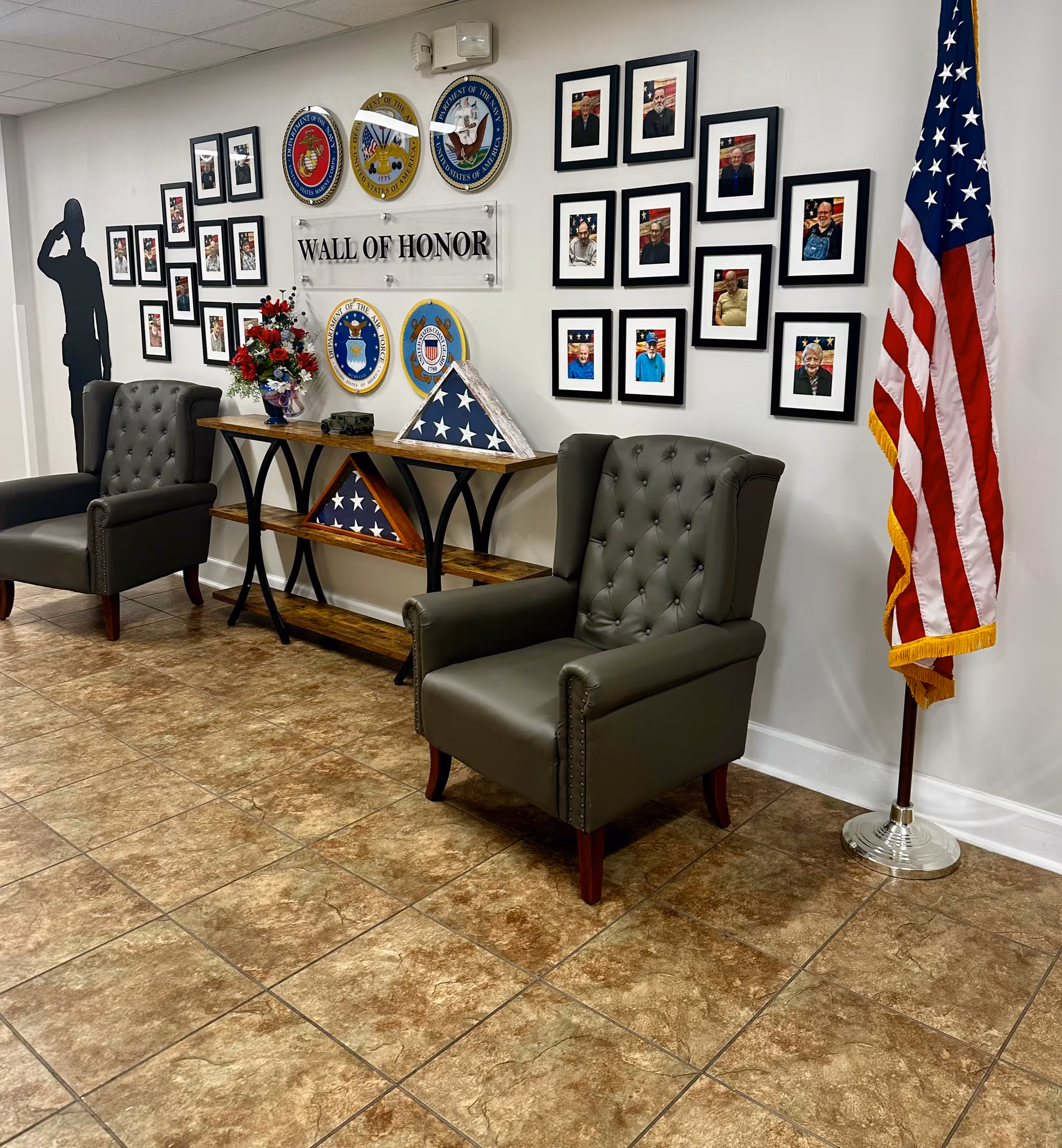 Interior view of a room with two dark green armchairs on a tiled floor. Between the chairs is a wooden console table holding folded American flags and a flower arrangement. On the wall behind the table is a 'Wall of Honor' display featuring military emblems and framed photographs of veterans. An American flag stands on a pole to the right side of the image.